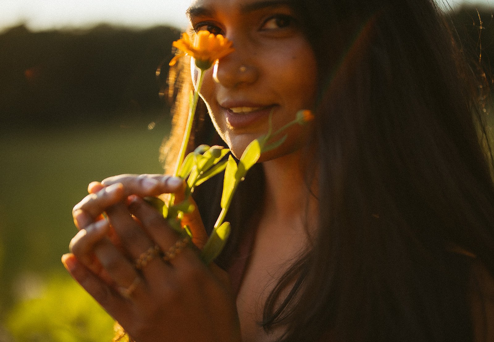 A woman with dark hair, holding yellow flowers close to her face, smiling gently in warm sunlight.