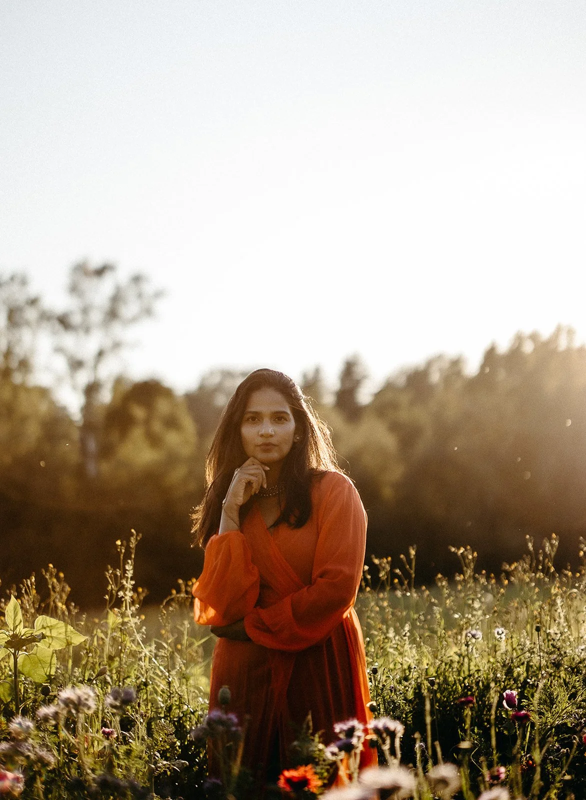 A woman in a red dress standing in a field of flowers at sunset.