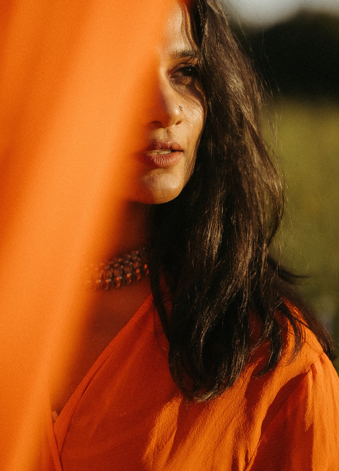 A woman with dark wavy hair, wearing orange clothing and a beaded necklace, partially obscured by an orange object in the foreground.