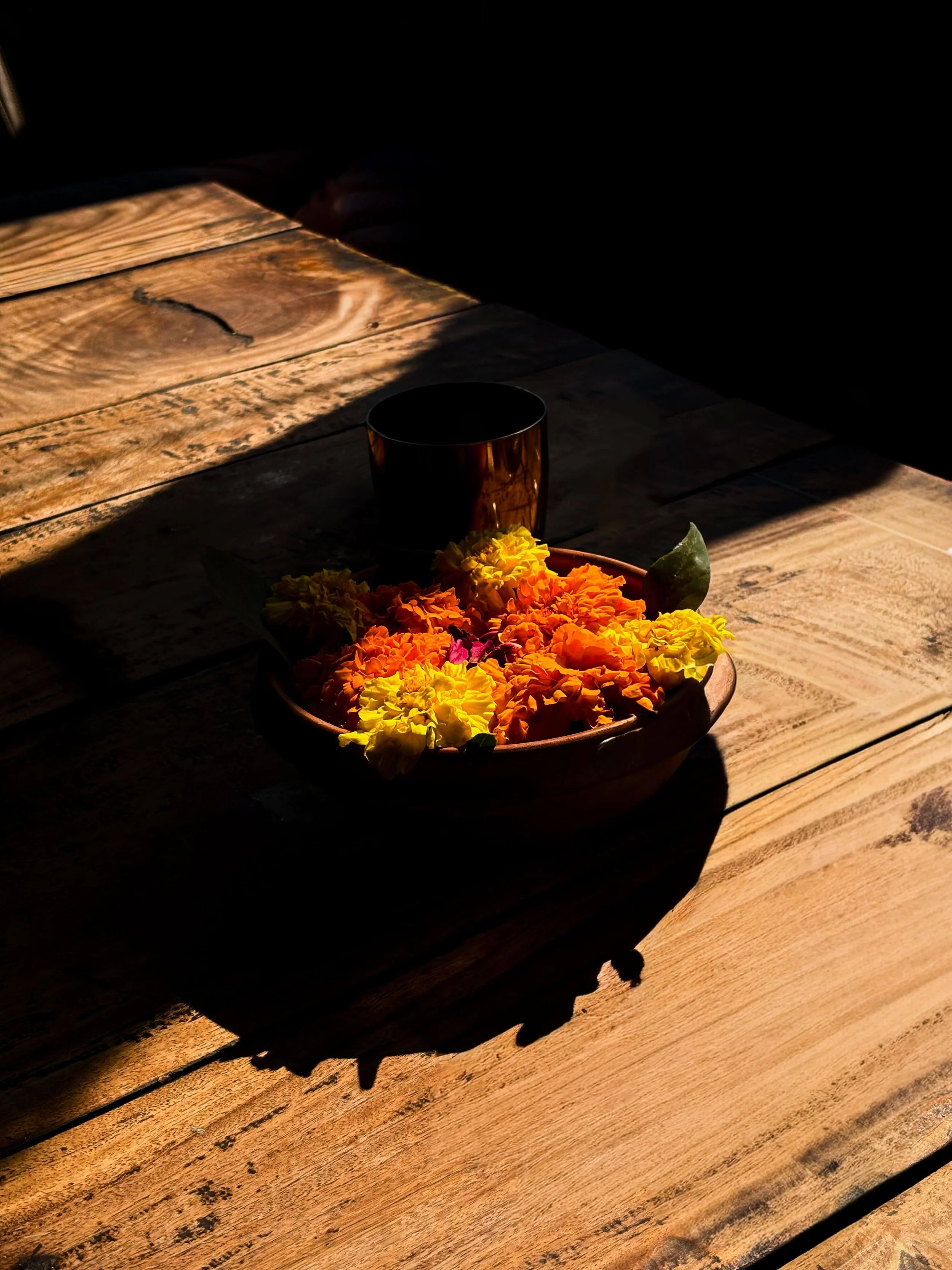 A wooden table with a flower arrangement in a bowl, a dark-colored cup, and a shadow cast across the surface.