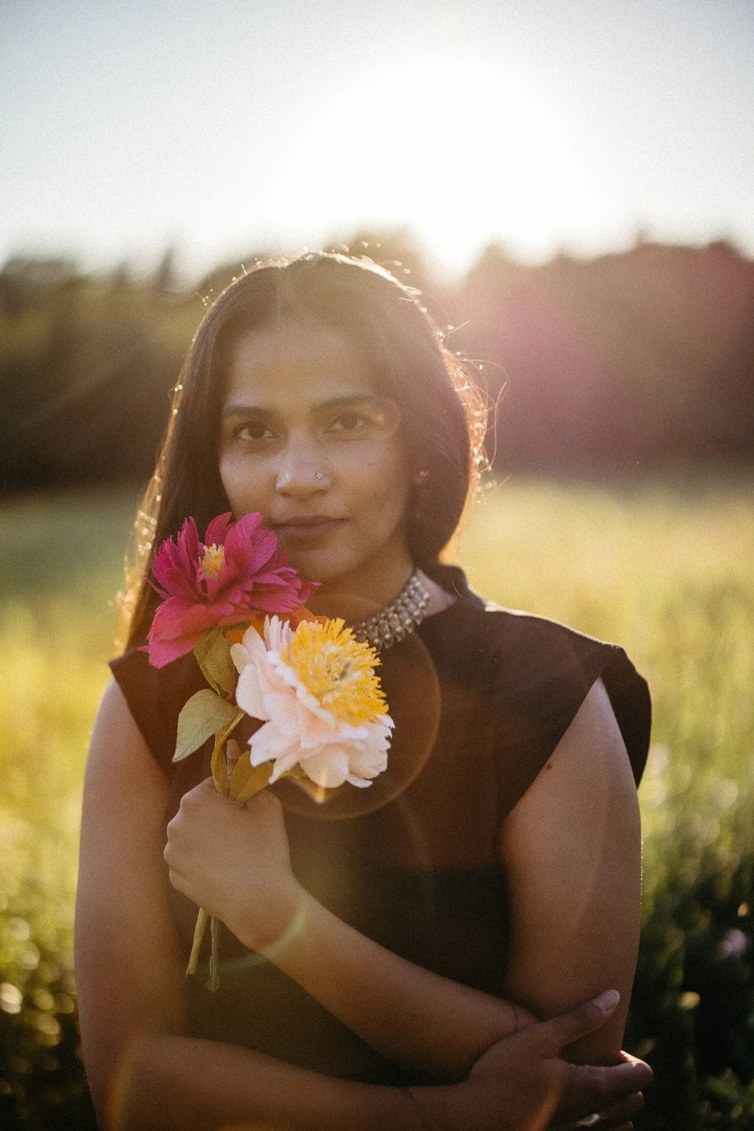 A woman holding a bouquet of pink, white, and yellow flowers stands outdoors at sunset, with sunlight behind her creating a warm glow.