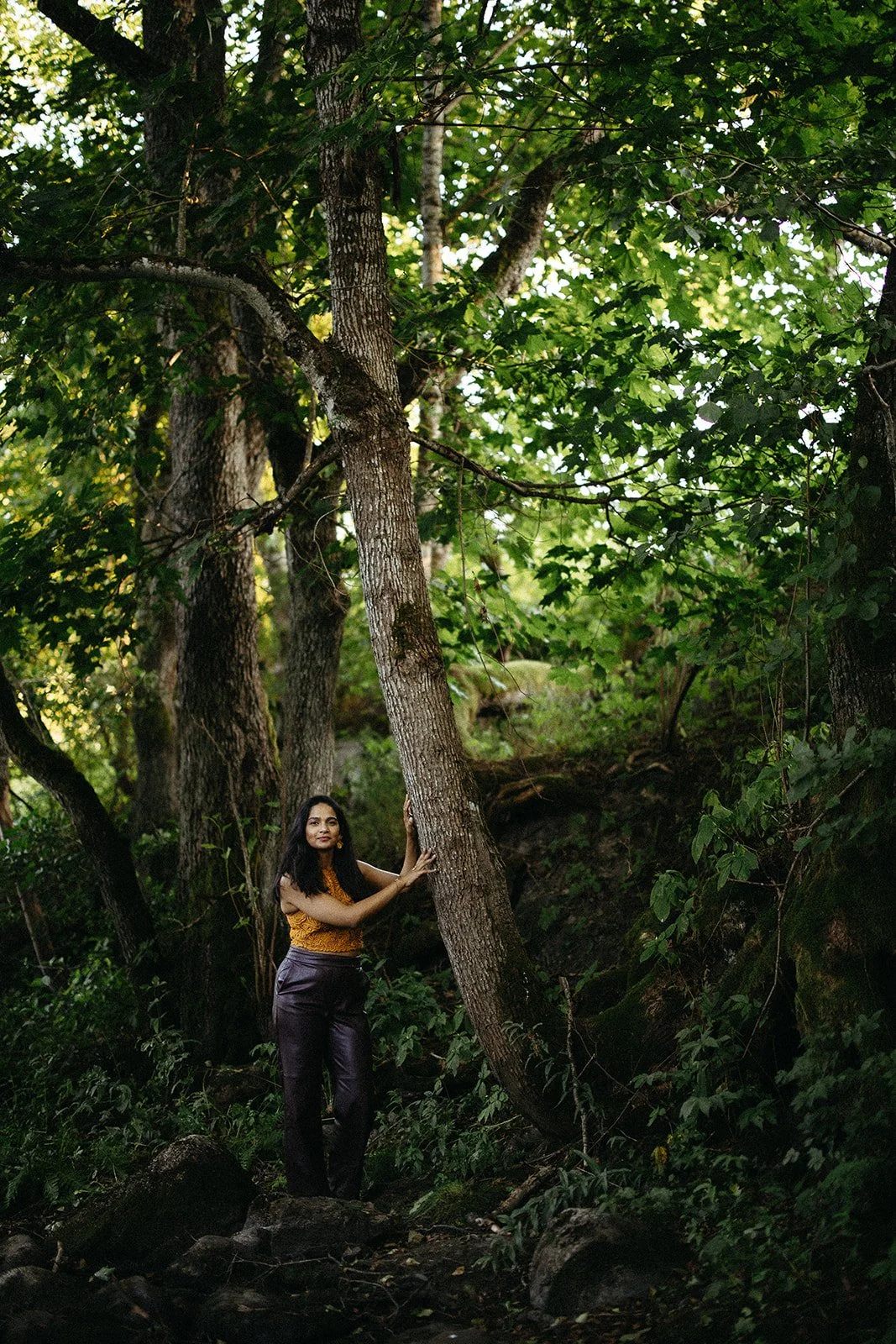 A woman in an orange top and dark pants standing in a forest, holding onto a tree trunk with green leaves and dense foliage around her.