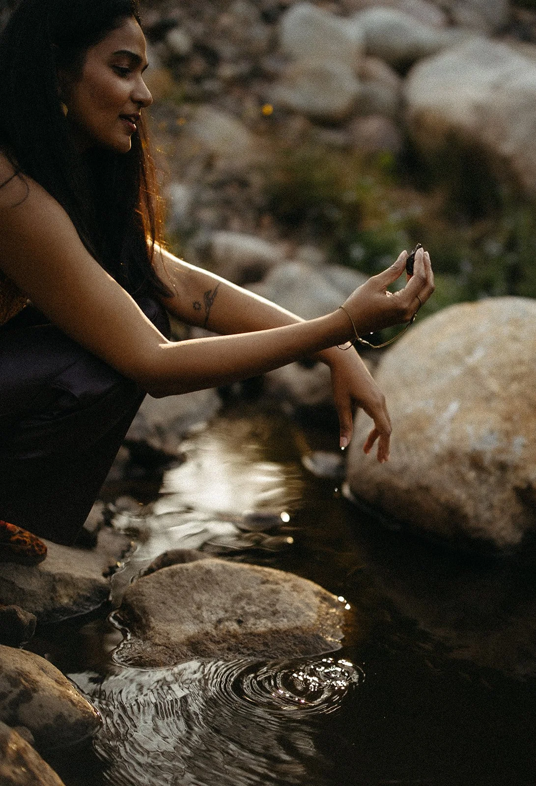 A woman with dark hair and tattoos on her arm sitting by a river, holding a small black object in her hand, with rocks surrounding her.