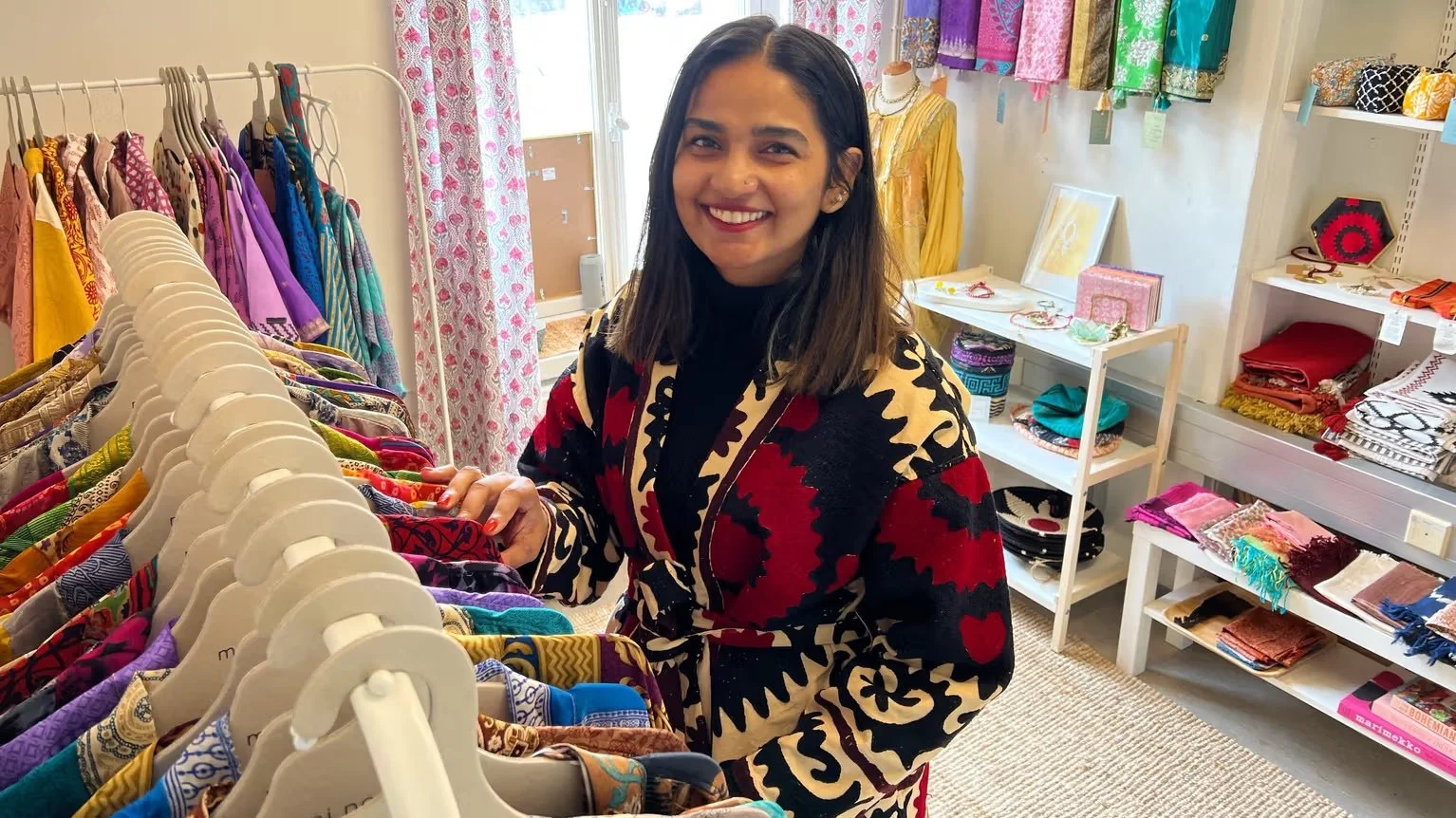 A smiling woman shopping in a clothing boutique, browsing through colorful scarves and garments on a rack.