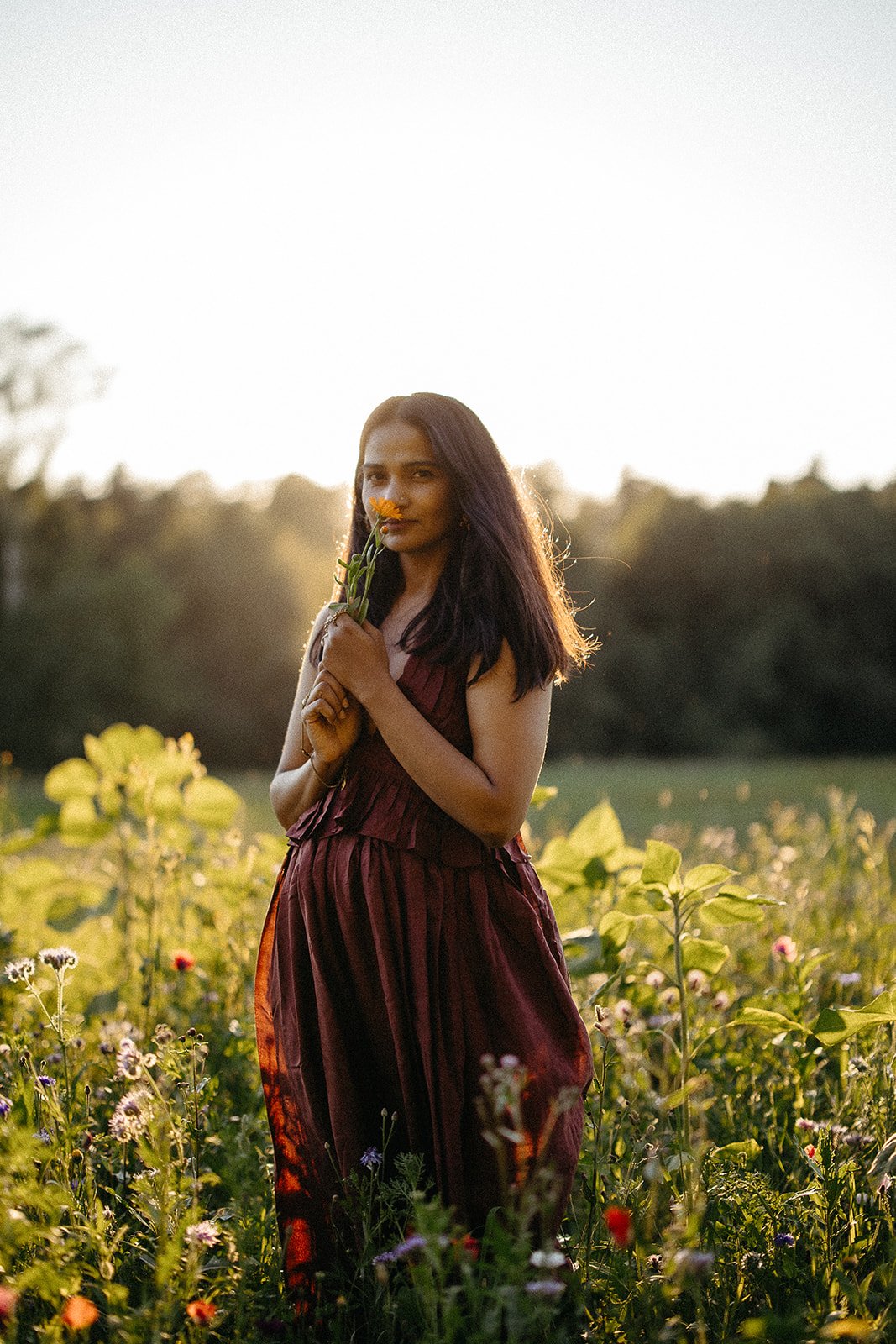 A woman in a maroon dress standing in a flower field holding and smelling a yellow flower at sunset.