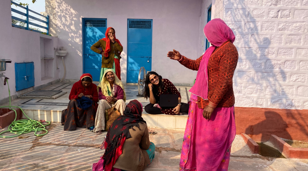 A group of women, some seated and some standing, are gathered outside near a building with a white wall and blue doors, engaged in conversation.