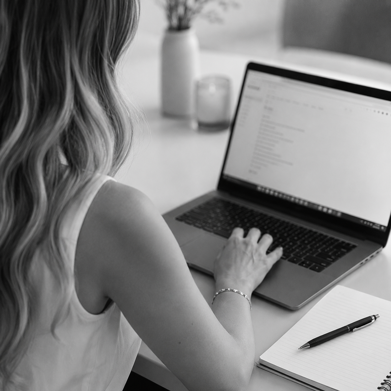A woman with wavy hair is working on a laptop at a desk, with a notebook and pen nearby, in a well-lit room with a vase and plants in the background.