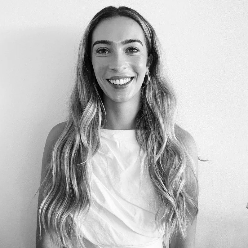 A smiling woman with long wavy hair, wearing a sleeveless top, standing against a plain wall.