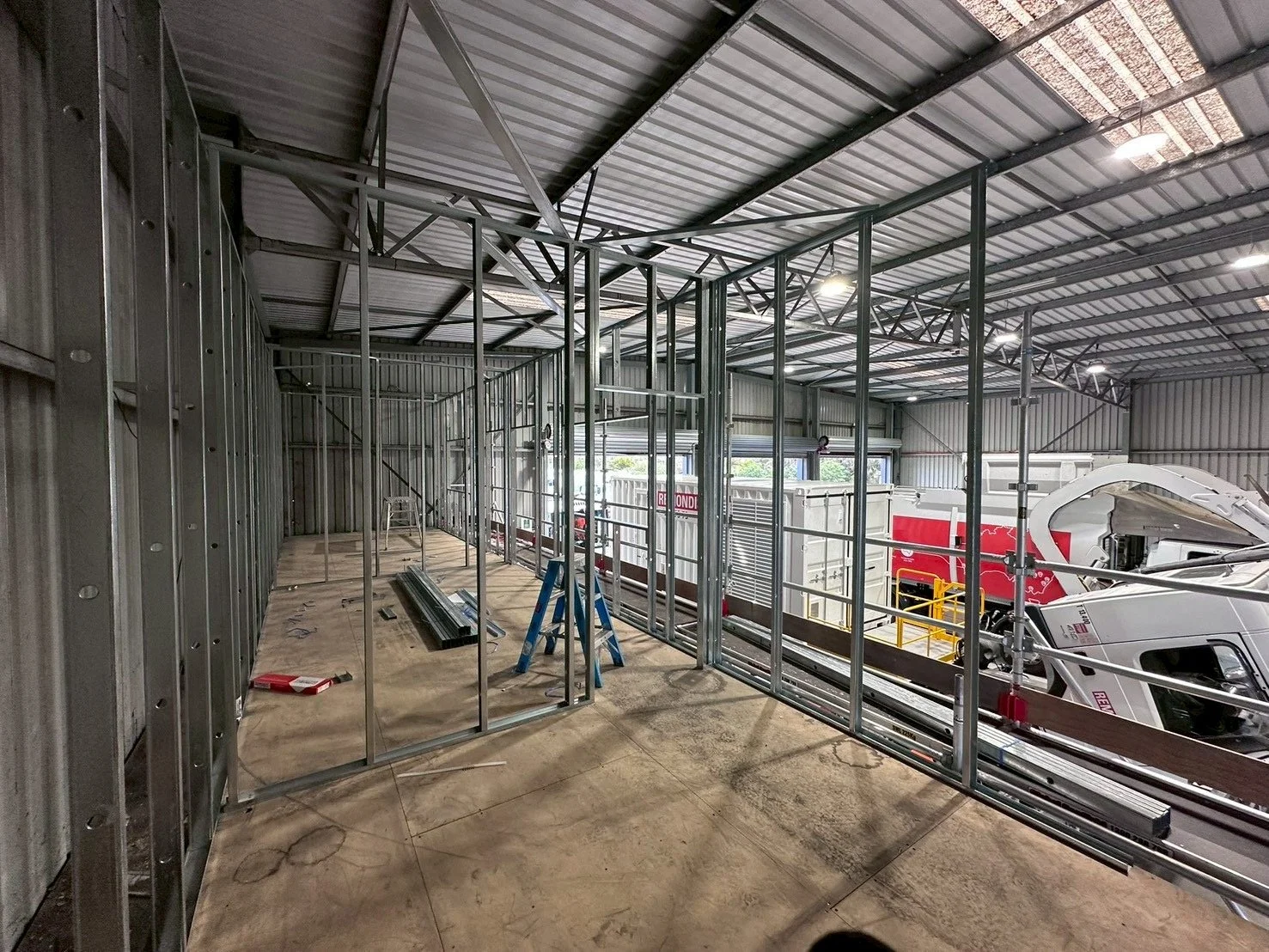 Construction of a building interior with metal framing, a ladder, and partially assembled walls. Garage doors and trucks are visible outside.