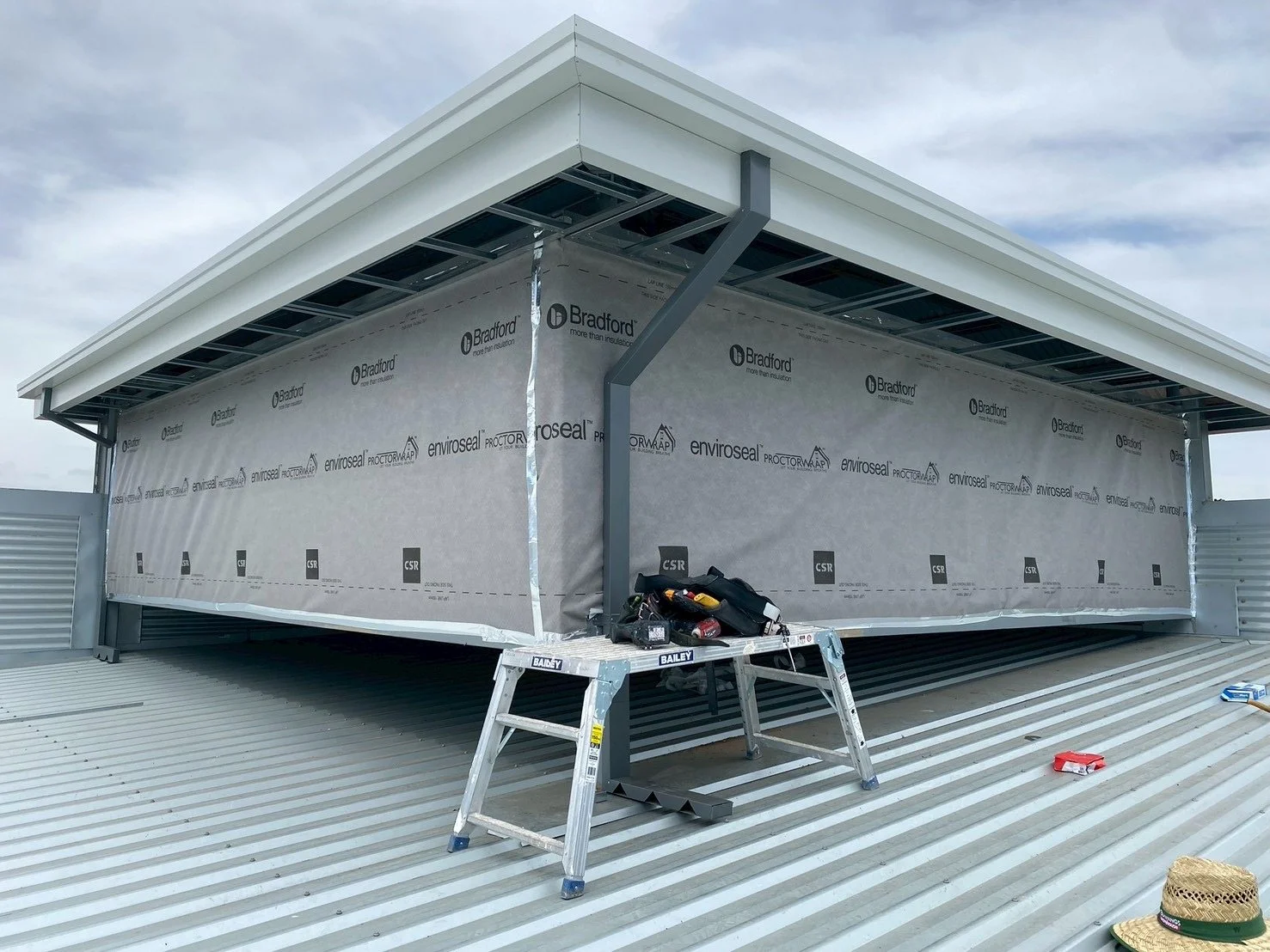 Construction work on a building roof with a partially installed wall wrapped in protective material, a silver step ladder, and various tools on the roof.