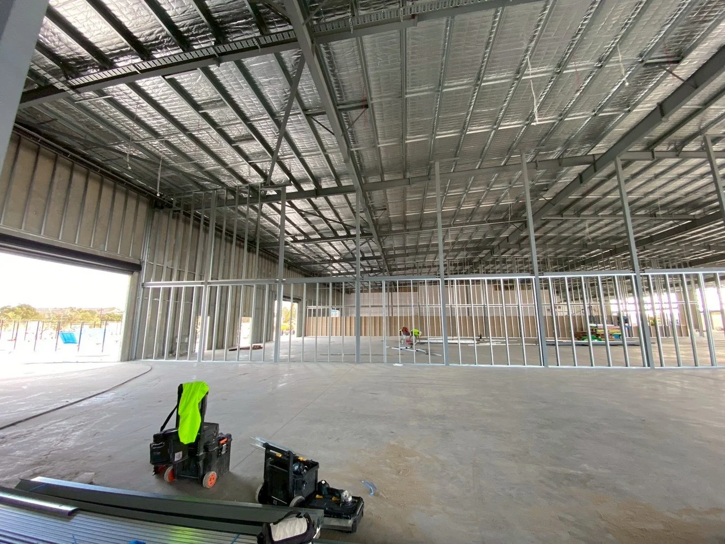 Construction site inside a building with metal framing and open wall spaces, tools and equipment are on the floor, and large open doorway with sunlight coming through.
