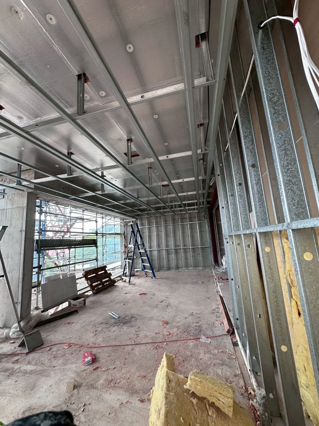 Construction site with metal framing, ladders, and insulation on the floor, indicating building interior under renovation.