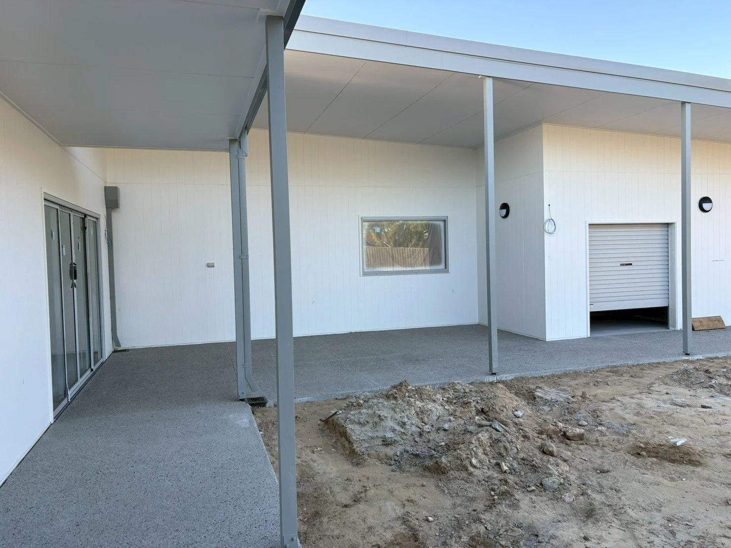 Corner of a modern house exterior with a covered patio, sliding glass doors, a small window, a closed garage door, and outdoor wall lights, with dirt and construction debris in the foreground.