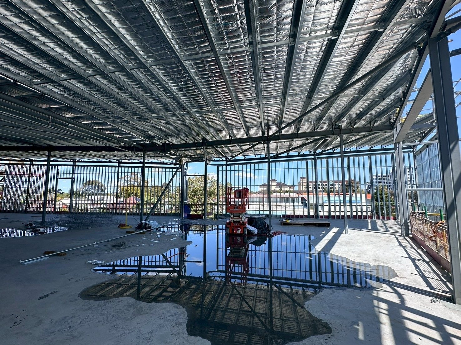 Construction site of a building with steel framework and reflective ceiling, showing a large puddle of water on concrete floor and city view through perimeter metal fencing.