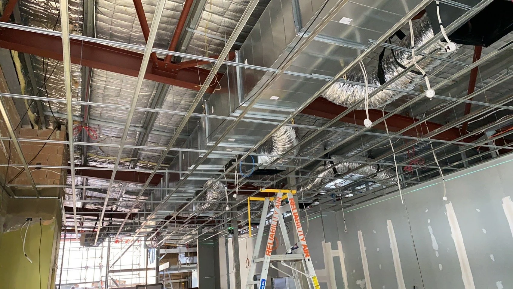 Construction site interior with exposed ceiling framework, ductwork, and electrical wiring, with a step ladder in the foreground.