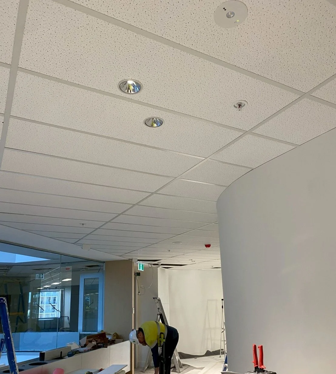 Interior construction or renovation area with a worker in a yellow jacket and white helmet working on the floor, ladders, construction tools, and a curved white wall.