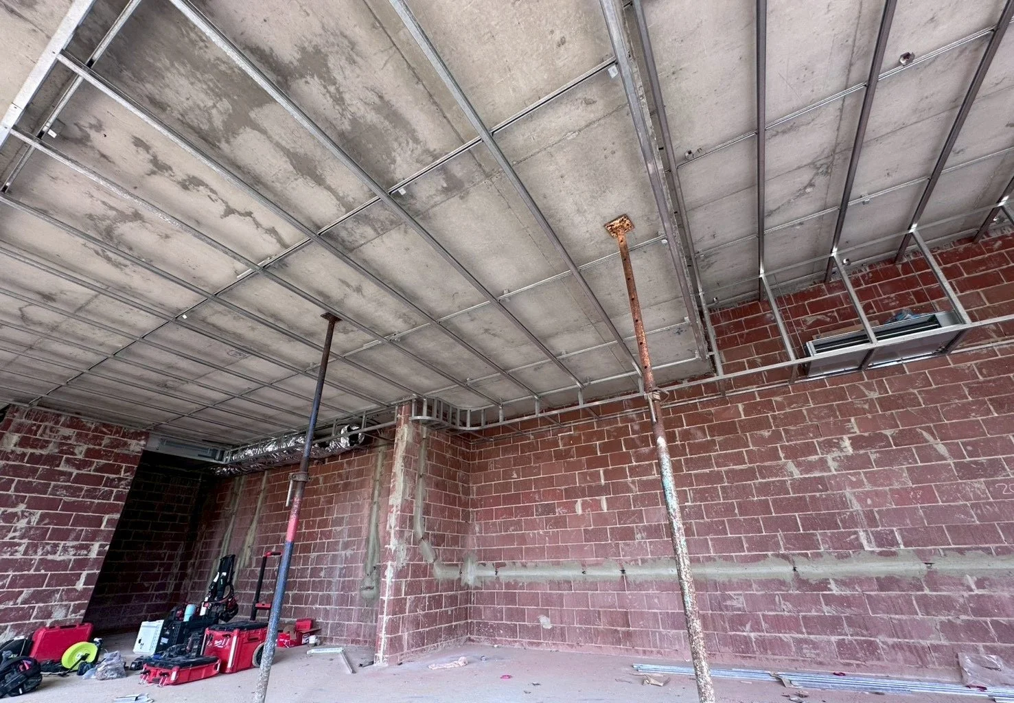 Construction site with exposed brick walls, metal ceiling framing, and support poles. Construction tools and equipment are on the floor.