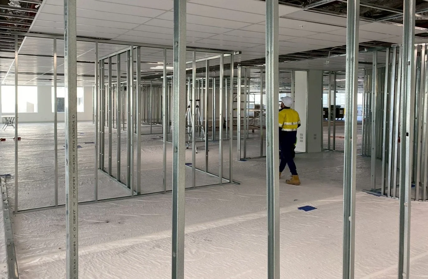 Construction worker in yellow safety vest and white helmet walking through an unfinished office space with metal wall frames and a suspended ceiling.