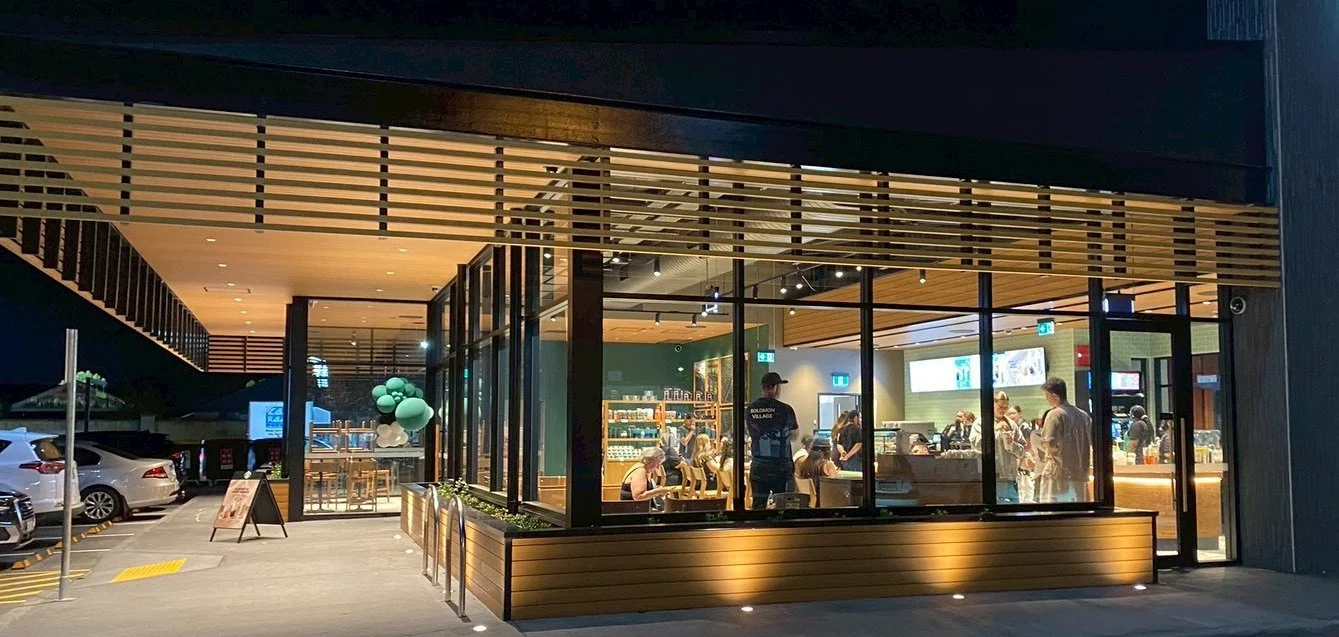 Night view of a modern glass-walled restaurant with busy indoor dining, outdoor parking with cars, and balloons decorating the entrance.