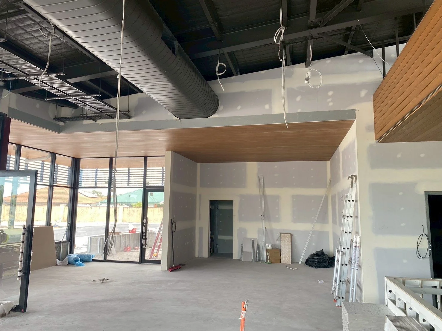 Interior of a building under construction with unfinished walls, ceiling, and exposed ductwork. Construction tools, ladders, and materials are visible around the space.