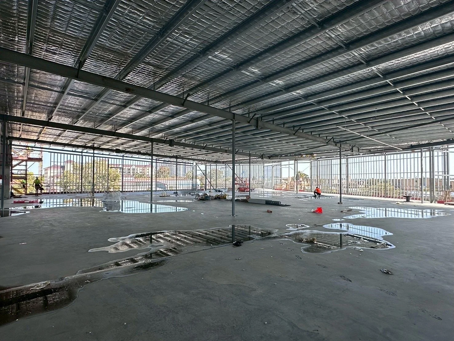 An open construction site with a concrete floor and a metal framework ceiling. Puddles of water are scattered across the floor, and there are construction workers in the distance. The area is surrounded by metal fencing, and buildings can be seen outside.