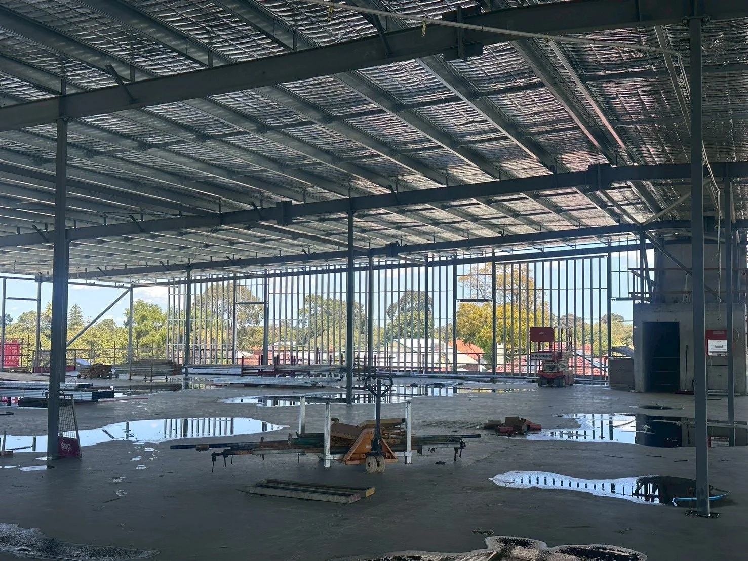 Interior view of a building under construction with steel framework, exposed insulation on the ceiling, large windows, and a concrete floor 