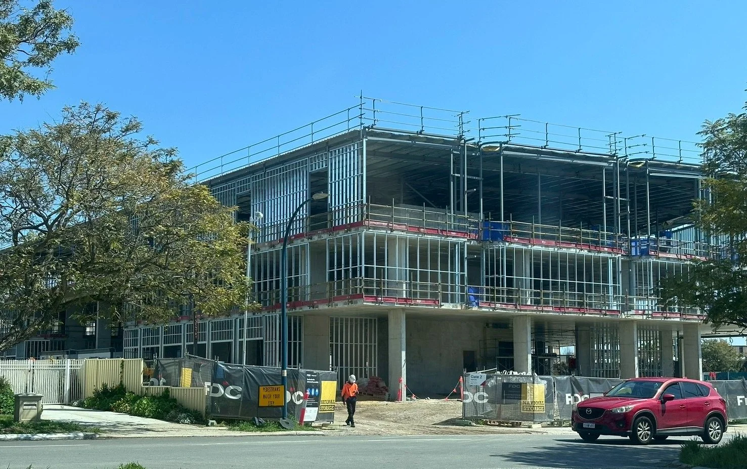 Multi-story building under construction with scaffolding and safety barriers, construction workers, cars parked nearby, trees, and a clear blue sky.