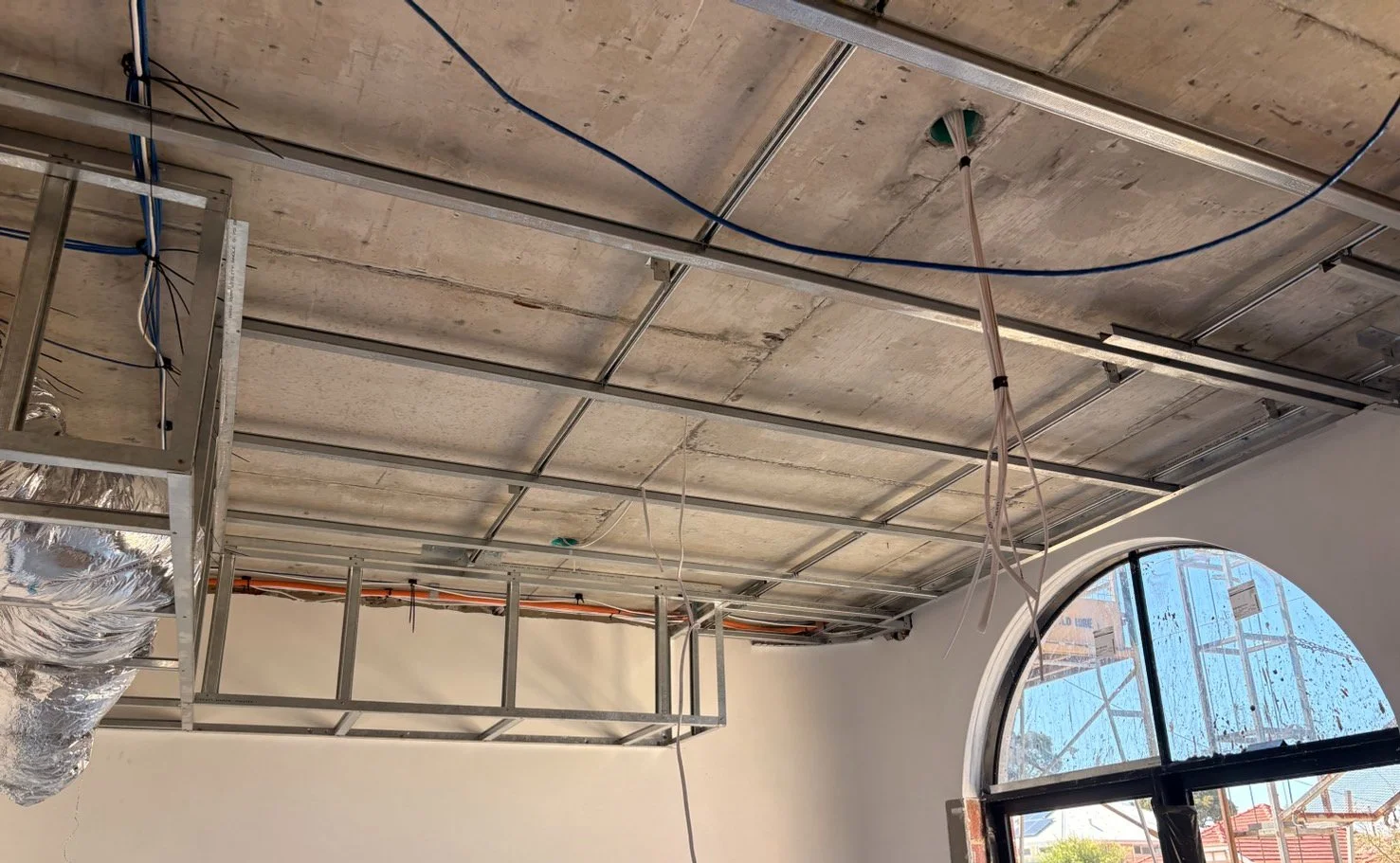 Interior of a building under construction with exposed ceiling framework, electrical wires hanging, and a large arched window showing scaffolding outside.