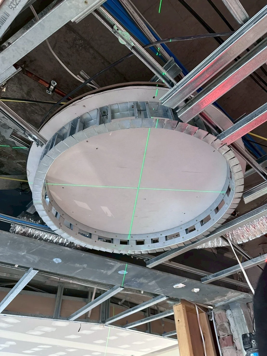 Construction site with a circular structure suspended from the ceiling, with visible metal framing, wooden elements, and green laser level lines.