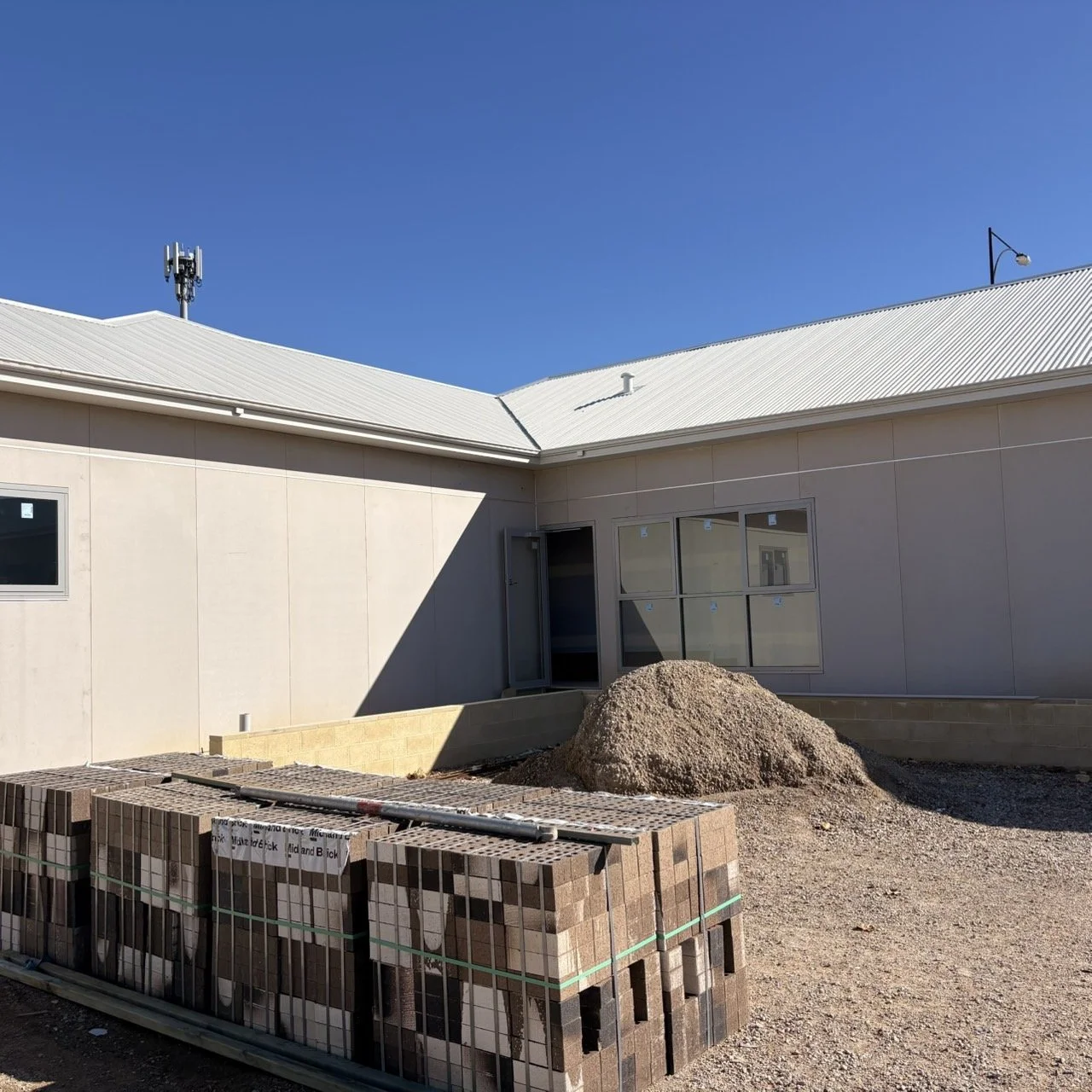 Construction site of a modern building with beige walls and a white metal roof, with bricks and a pile of dirt in the foreground, and a clear blue sky.