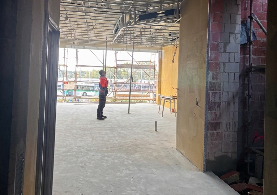 A person standing inside an unfinished building under construction, looking out through the open front. The construction site has scaffolding, construction materials, and unfinished walls.