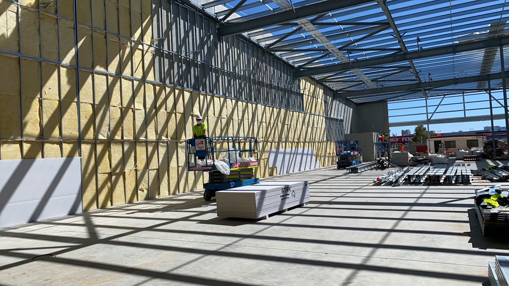 Construction site with workers and equipment inside a building under construction. Sunny day with shadows cast by a metal roof structure.