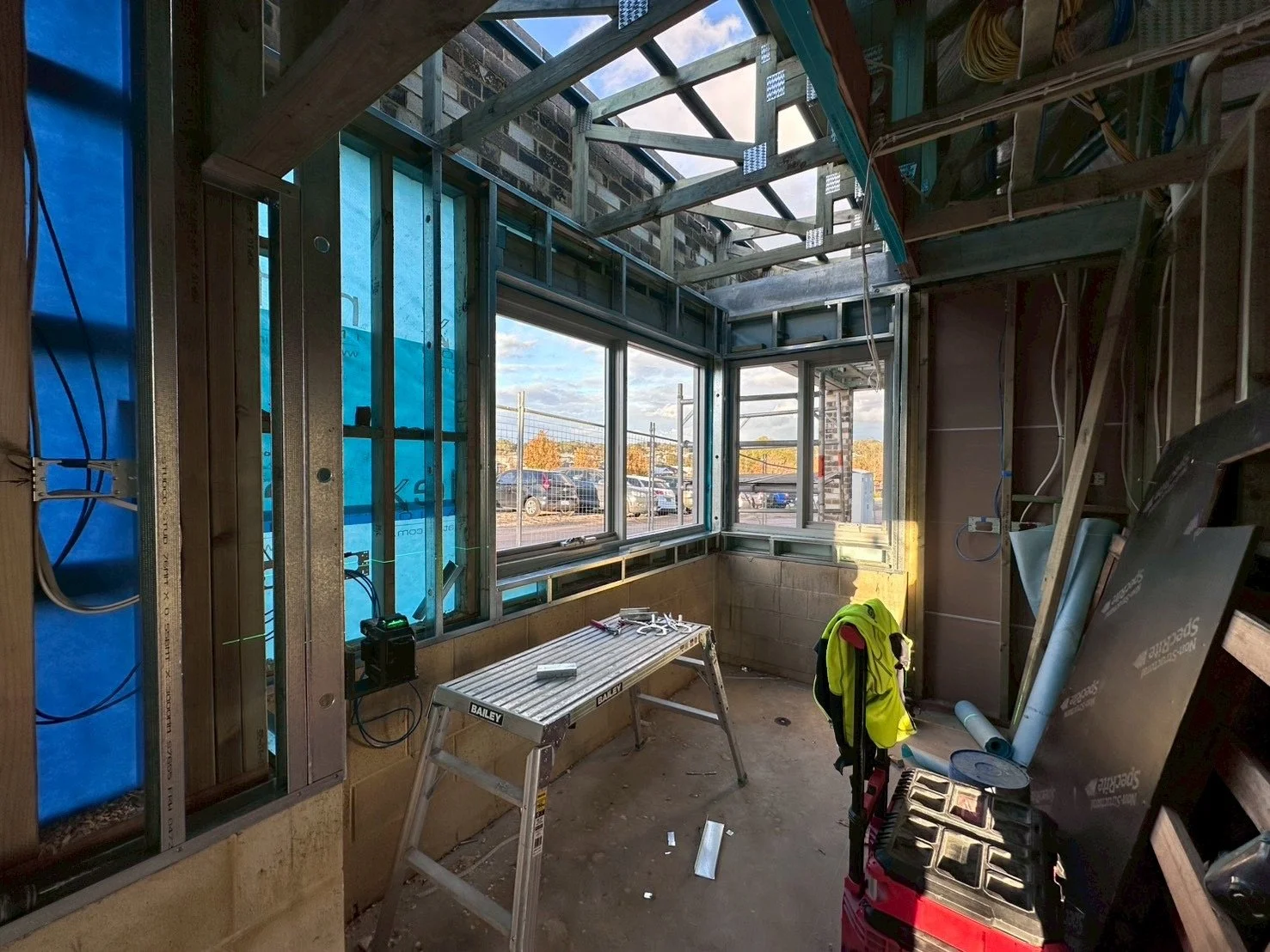 Construction site showing a partially built room with framed walls, large windows, and construction tools and materials inside.