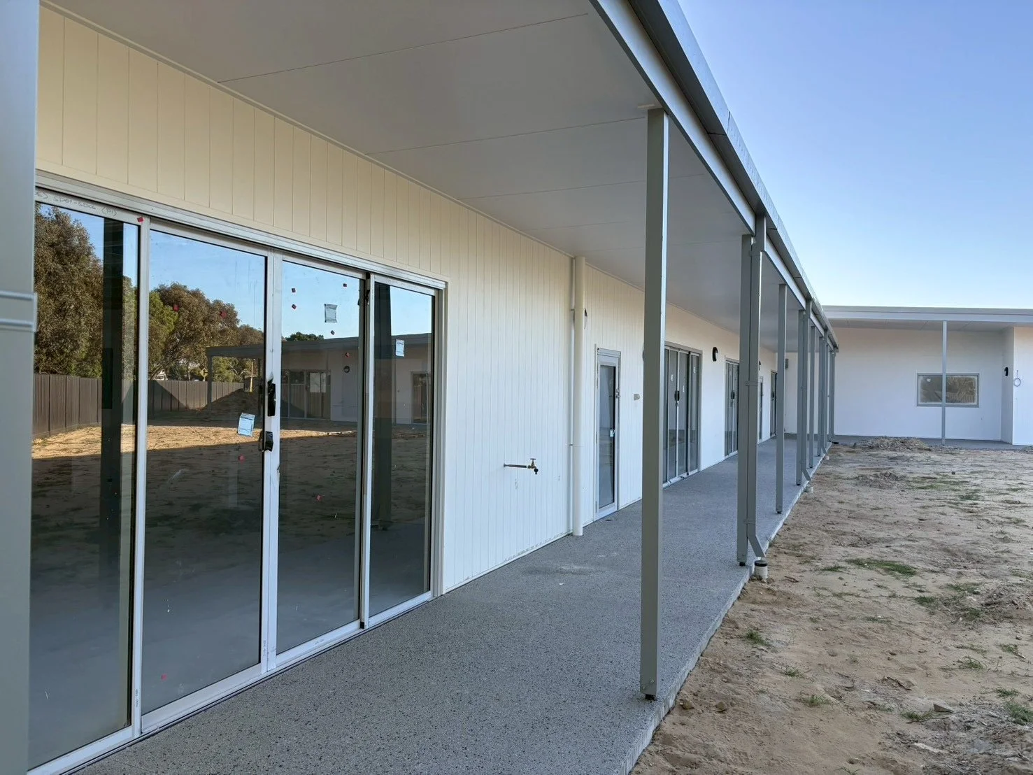 Newly constructed building with sliding glass doors, white exterior walls, and a covered walkway supported by metal columns. The ground around the building is bare dirt.