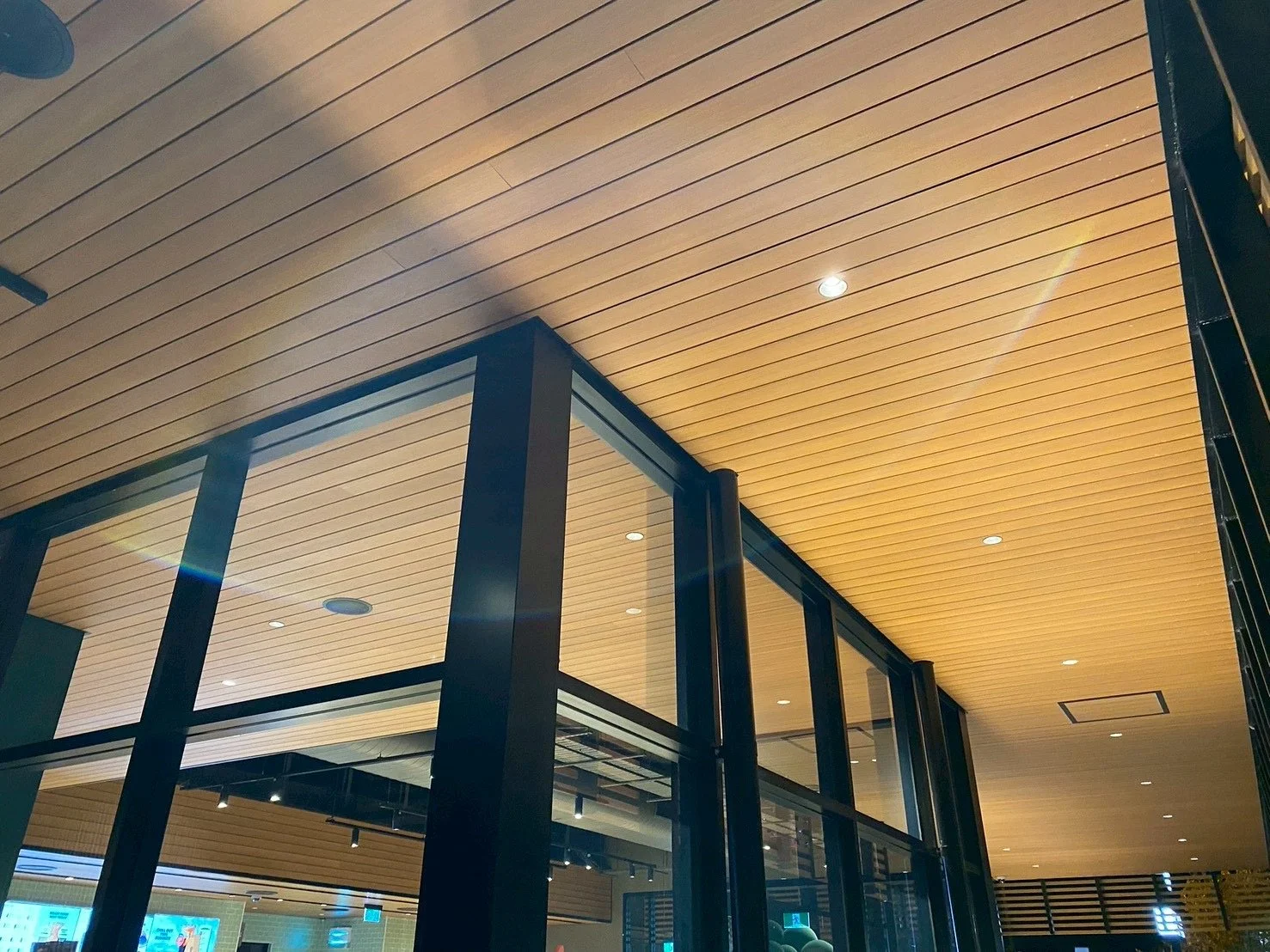 Interior view of a modern building with a wooden ceiling, glass walls, and black structural supports.