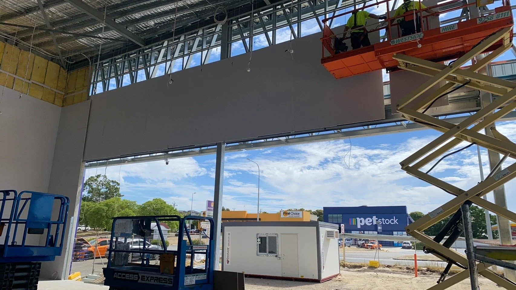 Construction workers on a scissor lift installing a large wall panel in a commercial building under construction, with visible steel framework and large windows showing a parking lot and retail stores in the background.