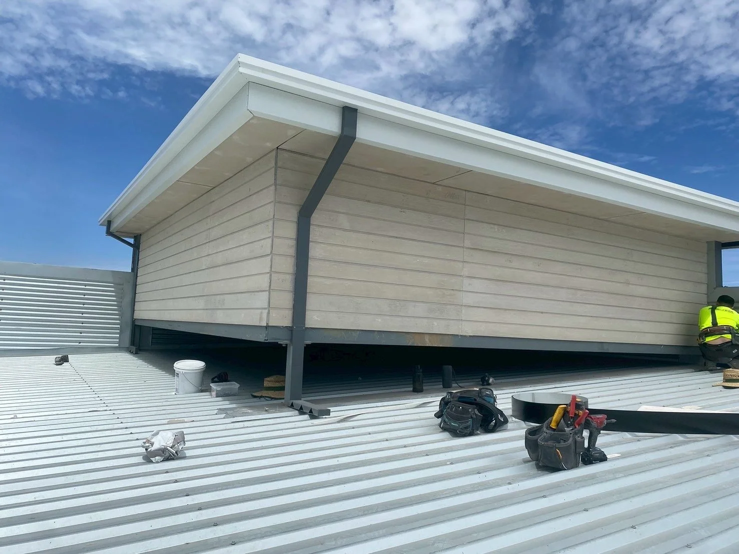 Building under construction with a worker, tools, and materials on a metal roof, sky with clouds in the background.