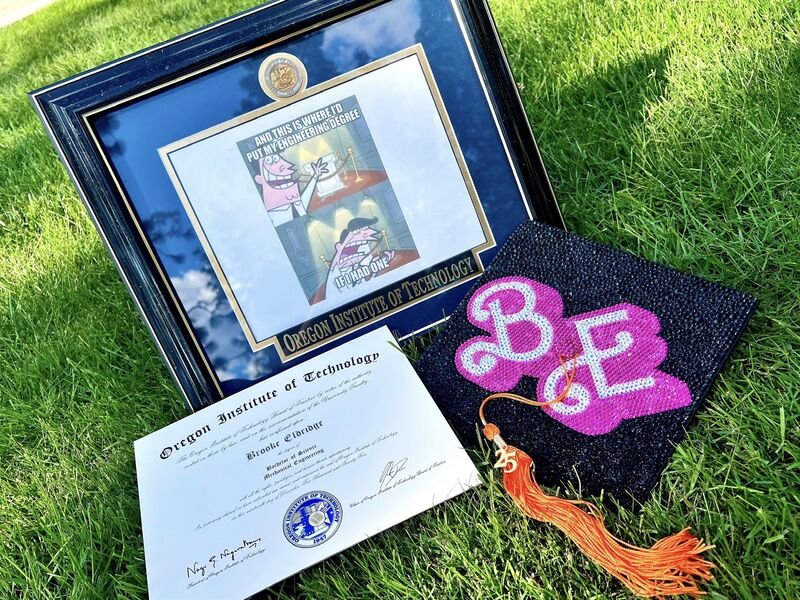 Graduation diploma from Oregon Institute of Technology, a decorated graduate cap with pink and white initials, and a framed cartoon comic about engineering, all displayed on green grass.