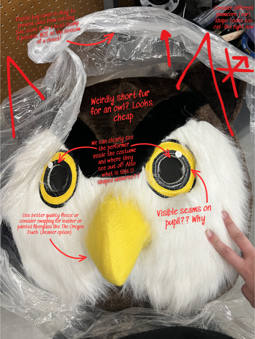 A craft project of an owl mask in progress, with annotations in red text and arrows pointing to different parts of the mask. The mask has a yellow beak, furry white face, black and yellow eyes with seams, and a brown furry owl face. A clear plastic bag cover is draped over the mask to prevent dust, and additional supplies can be seen in the background.