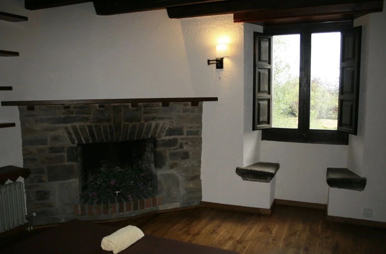 Interior de una habitación con chimenea de piedra, ventana con contraventanas de madera abiertas y paredes blancas. Sofá con una toalla sobre él y suelo de madera.