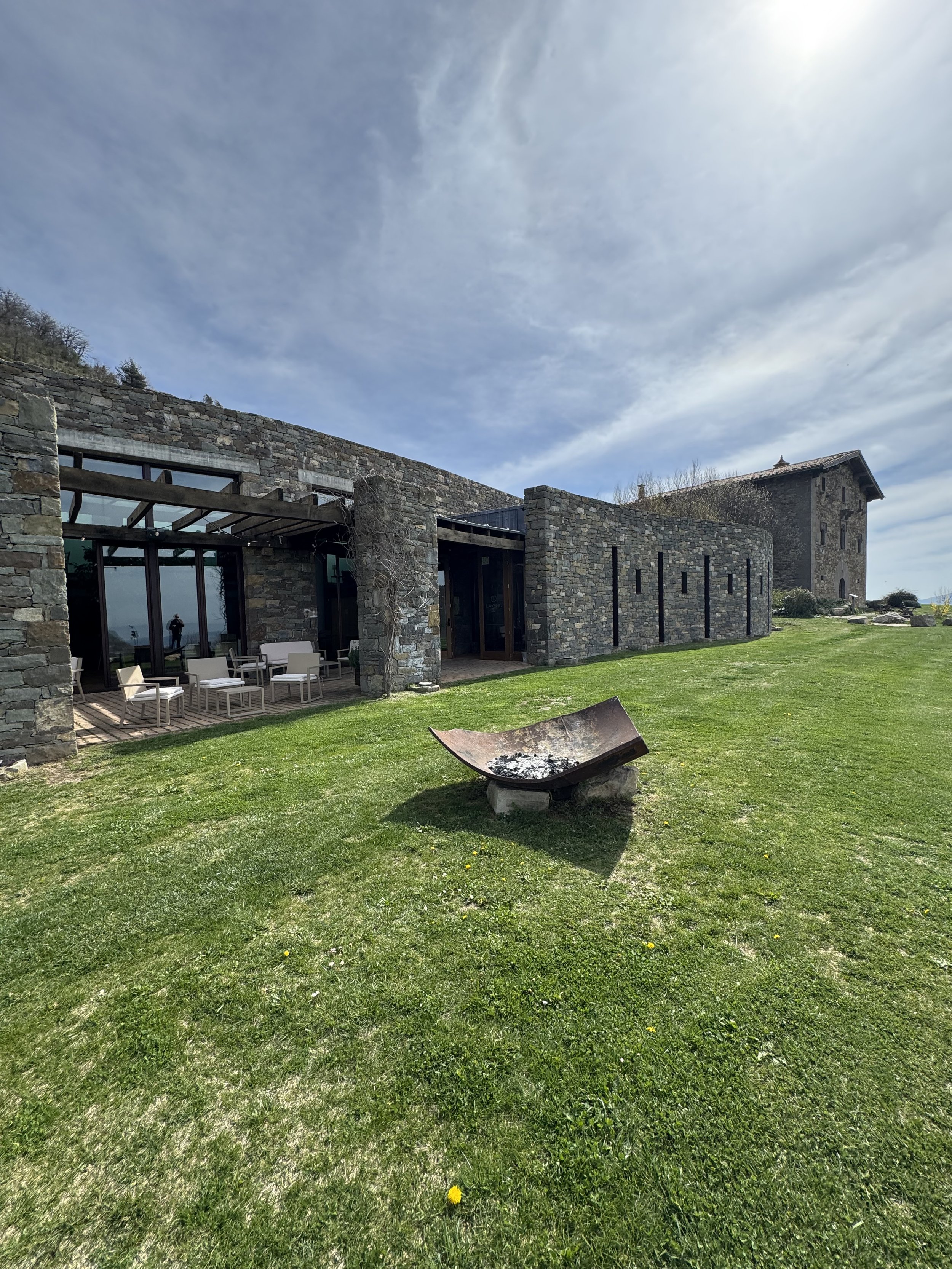 Edificio de piedra con áreas abiertas y mobiliario de exterior, rodeado de césped y un cielo con nubes.