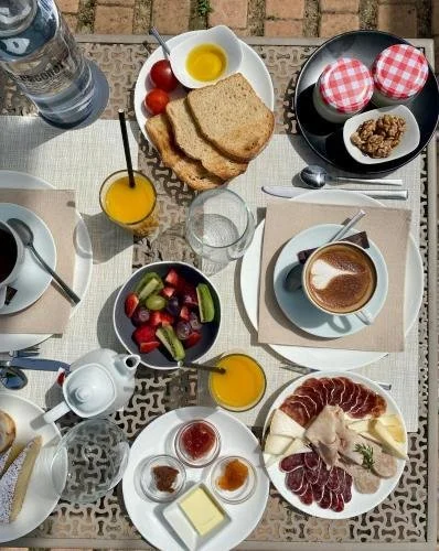 Desayuno completo con pan, fruta, huevos, embutidos, queso, yogurt, jugo, agua y café en una mesa al aire libre.