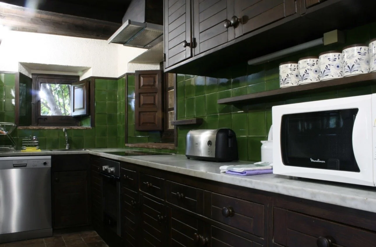 Cocina con gabinetes de madera, pared de azulejos verdes, electrodomésticos como microondas, tostadora, y lavavajillas, y una ventana abierta con vista a árboles.