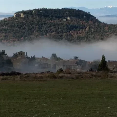 Vista de una colina o montaña con vegetación, neblina que cubre la zona baja y casas en primer plano.