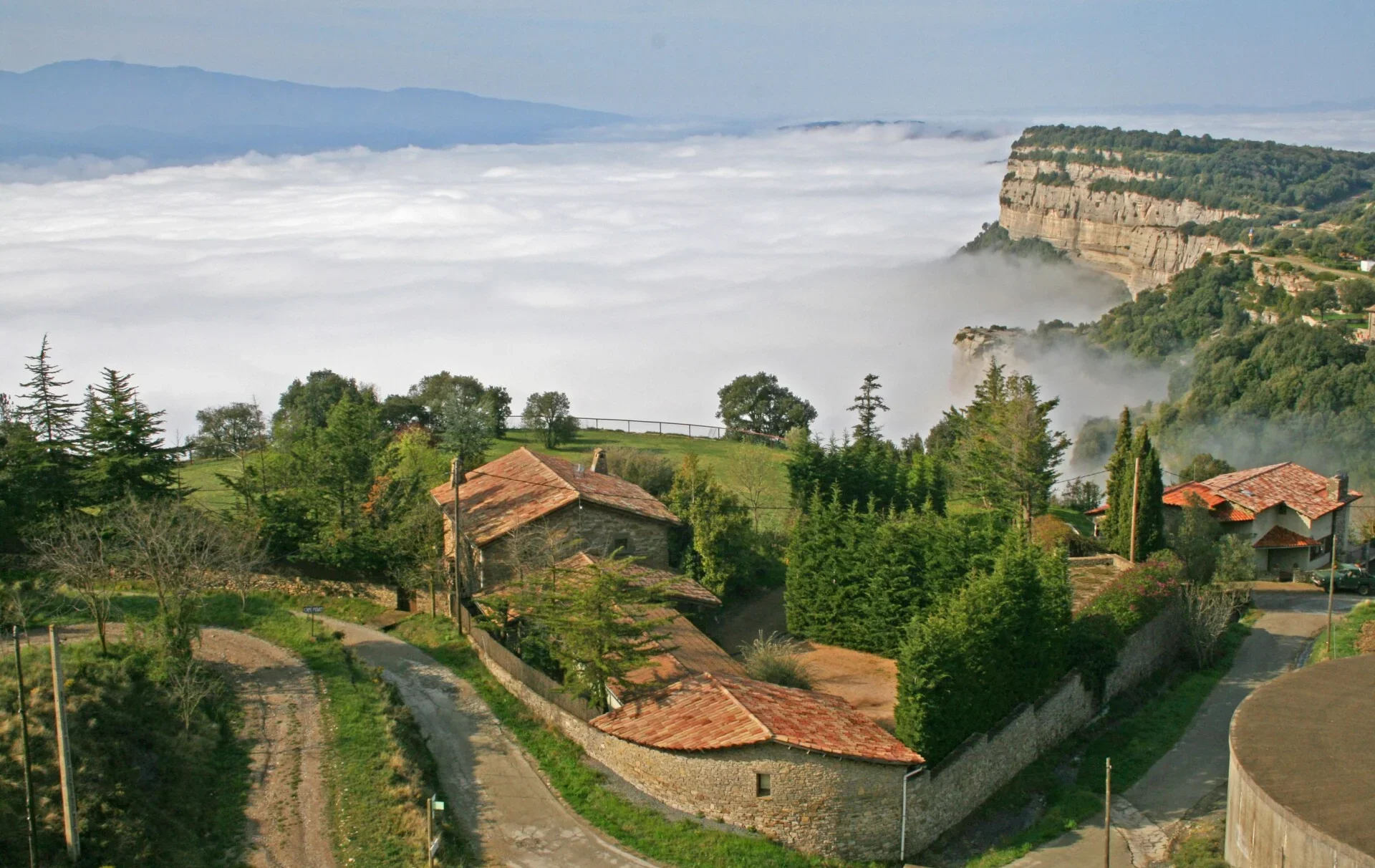 Paisaje de colinas verdes con casas de techos de tejas rojas y una neblina que cubre el valle, con acantilados y montañas al fondo.