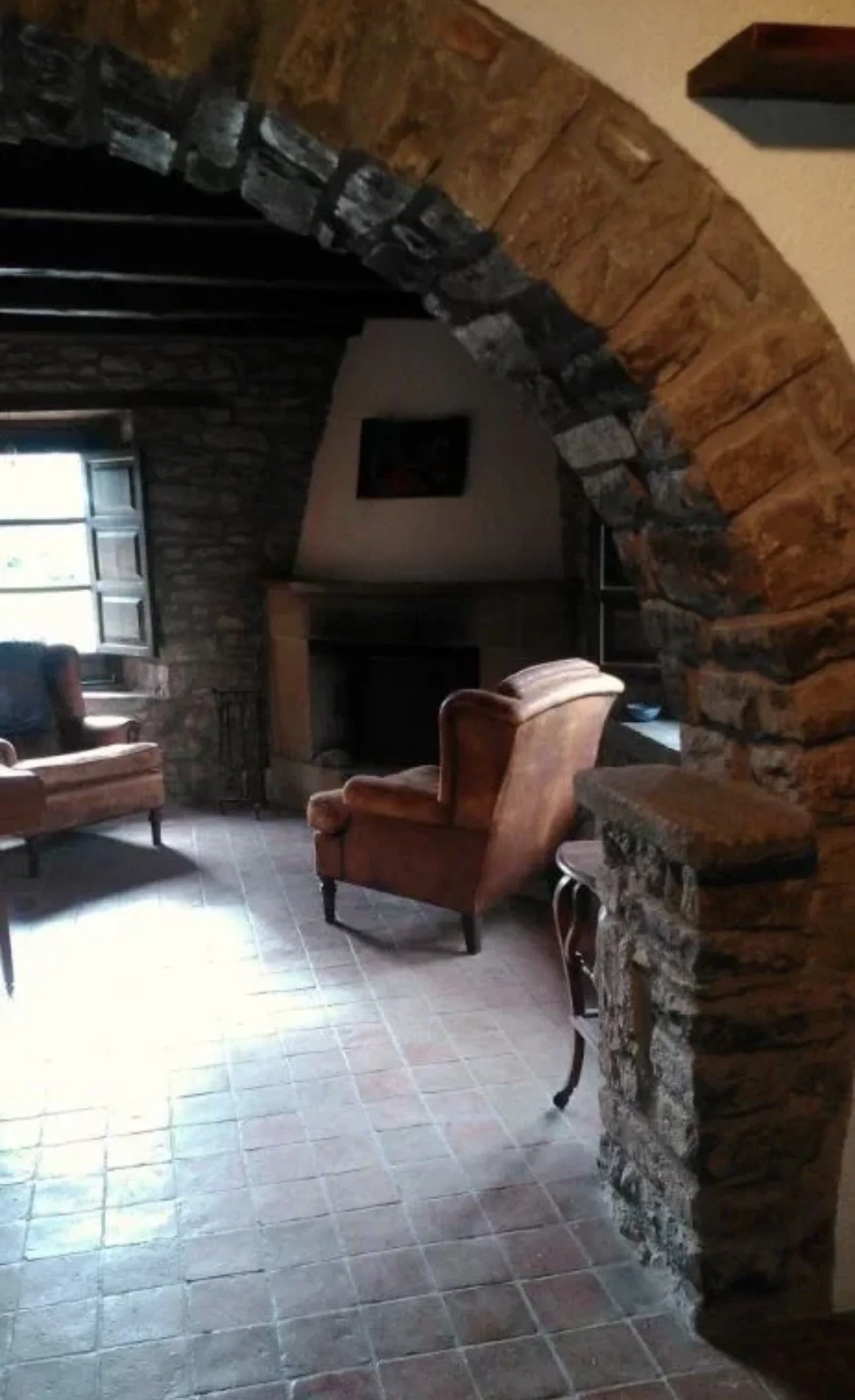 Interiores de una sala con paredes de piedra, sillones de cuero y una chimenea de piedra, con una ventana y cuadros en la pared, iluminación cálida.