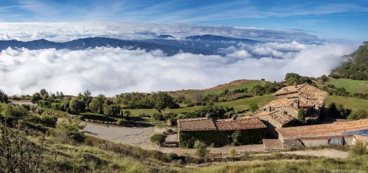 Vista de un pueblo en una zona montañosa con casas de tejado de teja y árboles verdes, con nubes y montañas al fondo.