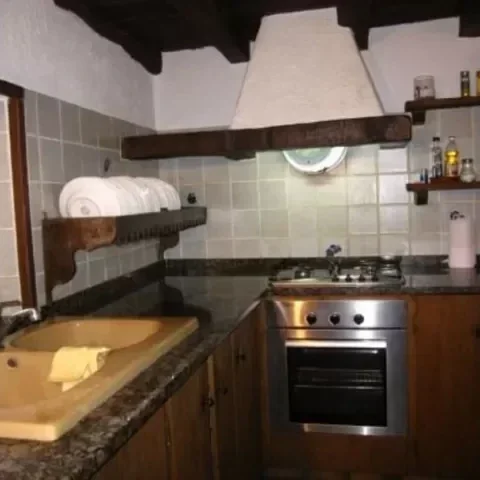 Cocina rústica con estufa de acero inoxidable, encimera de piedra, lavabo de cerámica, estantes de madera con utensilios y un campana extractora de madera en la pared.