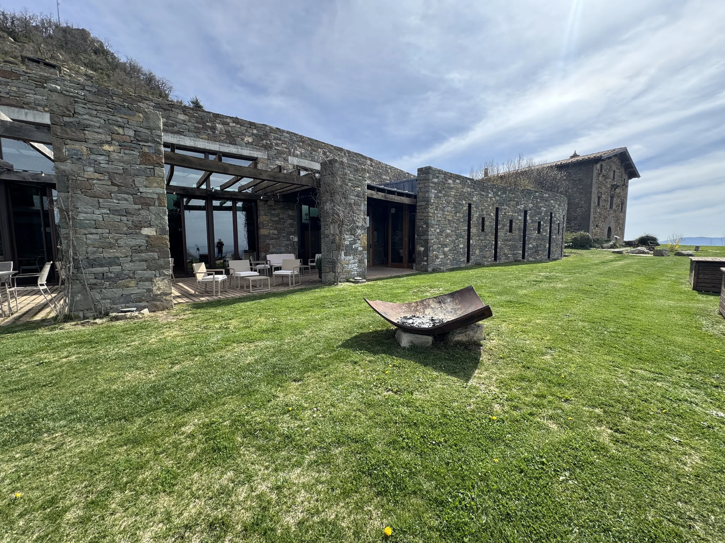 Edificio de piedra con ventanas grandes y mobiliario exterior en un campo verde, bajo un cielo nublado.