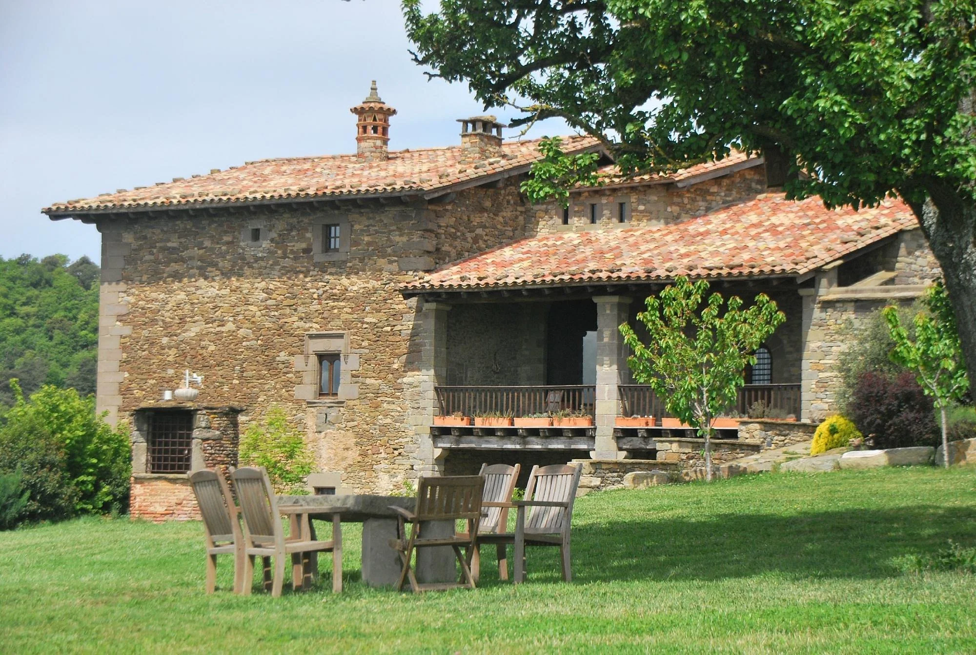 Casa de piedra con tejado de tejas rojas, rodeada de árboles y jardín, con mesas y sillas de madera en el césped.
